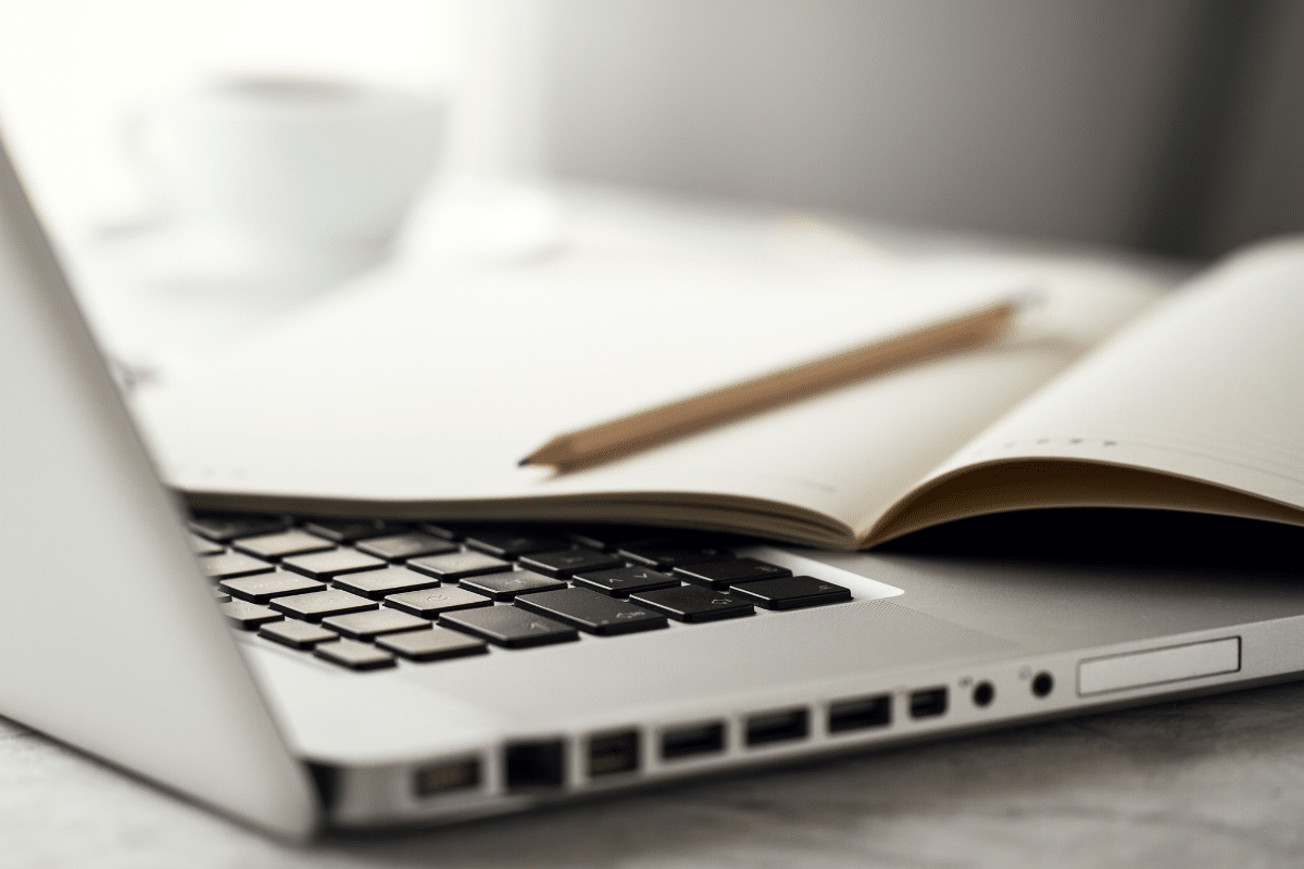 Notebook prateado com teclado preto, caderno aberto com lápis de madeira e xícara branca desfocada ao fundo