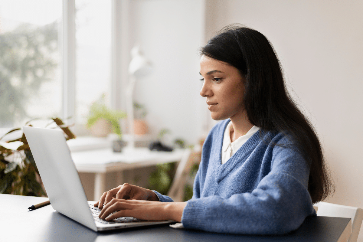 Mulher de cabelo escuro e suéter azul digita em laptop prata em ambiente iluminado com plantas ao fundo