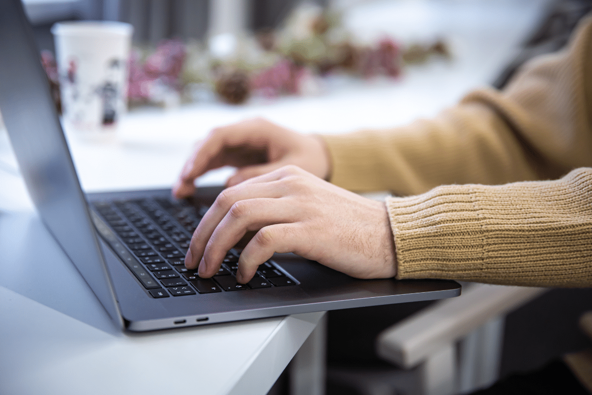 Mãos de uma pessoa digitando em um notebook sobre mesa branca com copo ao fundo
