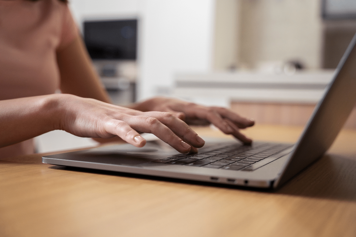 Jovem com camiseta rosa digitando em notebook sobre mesa de madeira em sala clara