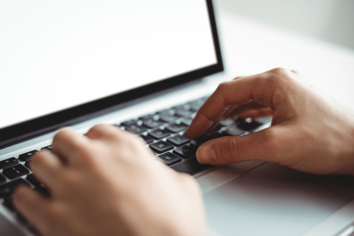 Duas mãos digitando em teclado preto de notebook prateado com tela em branco, fundo claro neutro