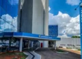 Imagem da fachada do INSS, mostrando o prédio com logo e nome 'Previdência Social' em destaque. A foto tem um céu claro e nuvens ao fundo, com elementos do edifício refletindo.