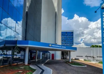 Imagem da fachada do INSS, mostrando o prédio com logo e nome 'Previdência Social' em destaque. A foto tem um céu claro e nuvens ao fundo, com elementos do edifício refletindo.