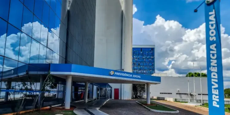 Imagem da fachada do INSS, mostrando o prédio com logo e nome 'Previdência Social' em destaque. A foto tem um céu claro e nuvens ao fundo, com elementos do edifício refletindo.