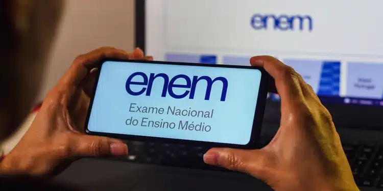 Mãos segurando smartphone com logo do ENEM na tela, notebook ao fundo exibindo página do participante
