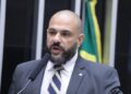 Deputado Sargento Portugal aparece com visual que adotou durante sua trajetória política: careca e barba. Enquanto esteve na Câmara dos Deputados, foi vice-líder do PSDB e vice-líder da Minoria. Veste terno cinza escuro com gravata listrada azul e branco, discursando ao microfone com bandeira brasileira desfocada ao fundo.