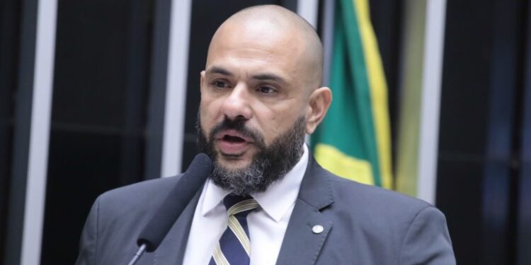 Deputado Sargento Portugal aparece com visual que adotou durante sua trajetória política: careca e barba. Enquanto esteve na Câmara dos Deputados, foi vice-líder do PSDB e vice-líder da Minoria. Veste terno cinza escuro com gravata listrada azul e branco, discursando ao microfone com bandeira brasileira desfocada ao fundo.