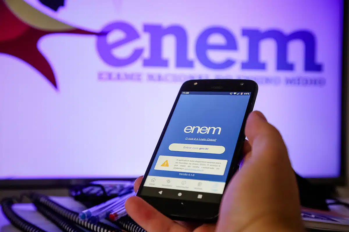 Mão segura celular Motorola com app ENEM aberto mostrando tela de login gov.br, monitor ao fundo exibe logo do exame