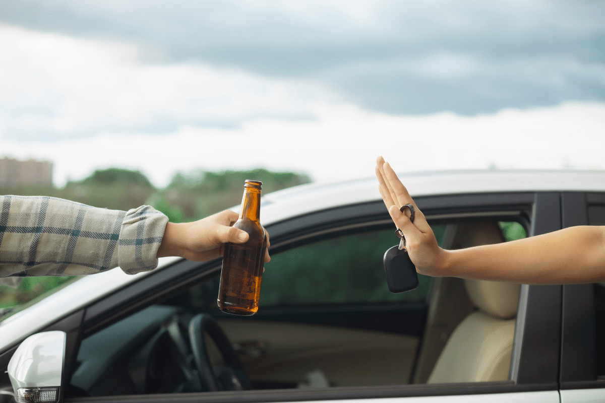 Mão erguida recusando garrafa de cerveja ao lado de carro cinza, segurando chaves do veículo
