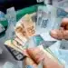 Notas de R$ 100 e R$ 50 sendo retiradas de um caixa eletrônico, representando o pagamento do INSS, com a liberação de R$ 8.475,55 para CPFs com finais de 0 a 9.