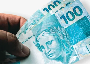 Notas de 100 reais sendo seguradas por uma mão, representando os pagamentos nos valores de R$ 811, R$ 1.351 e R$ 1.621 liberados em 15 de maio
