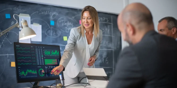 Profissional apresentando gráficos financeiros em tela para equipe durante reunião de estratégia no mercado financeiro.