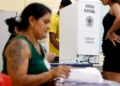 Imagem de mesários durante o processo eleitoral, com foco em uma cabine de votação e pessoas interagindo em uma seção eleitoral.