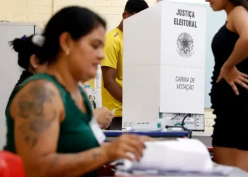 Imagem de mesários durante o processo eleitoral, com foco em uma cabine de votação e pessoas interagindo em uma seção eleitoral.