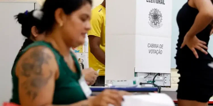 Imagem de mesários durante o processo eleitoral, com foco em uma cabine de votação e pessoas interagindo em uma seção eleitoral.