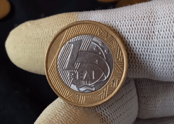 Close-up do reverso de uma moeda bimetálica brasileira de 1 Real, ano 2012. Uma mão com luvas brancas segura a peça bicolor, destacando o numeral '1', a inscrição 'REAL' e o ano '2012' sobre fundo texturizado.