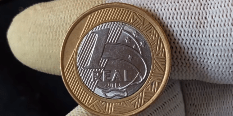 Close-up do reverso de uma moeda bimetálica brasileira de 1 Real, ano 2012. Uma mão com luvas brancas segura a peça bicolor, destacando o numeral '1', a inscrição 'REAL' e o ano '2012' sobre fundo texturizado.