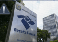 Placa de identificação da Superintendência da Receita Federal do Ministério da Fazenda em frente a edifício comercial, com ícone de alerta amarelo em destaque