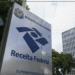 Placa de identificação da Superintendência da Receita Federal do Ministério da Fazenda em frente a edifício comercial, com ícone de alerta amarelo em destaque