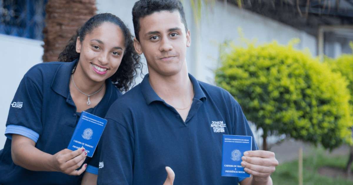 Jovem mulher e homem uniformizados sorriem ao mostrar carteira de trabalho azul, celebrando ingresso profissional