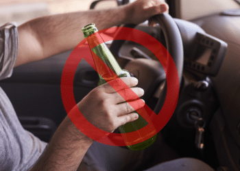 Mão masculina segura garrafa verde de cerveja atrás do volante com sinal vermelho de proibição sobreposto