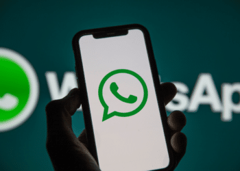 Mão segura celular preto mostrando ícone verde do WhatsApp, com logo ampliado ao fundo