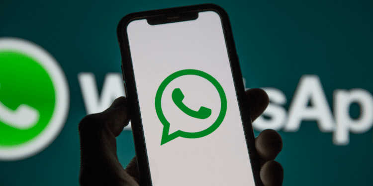 Mão segura celular preto mostrando ícone verde do WhatsApp, com logo ampliado ao fundo
