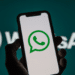 Mão segura celular preto mostrando ícone verde do WhatsApp, com logo ampliado ao fundo