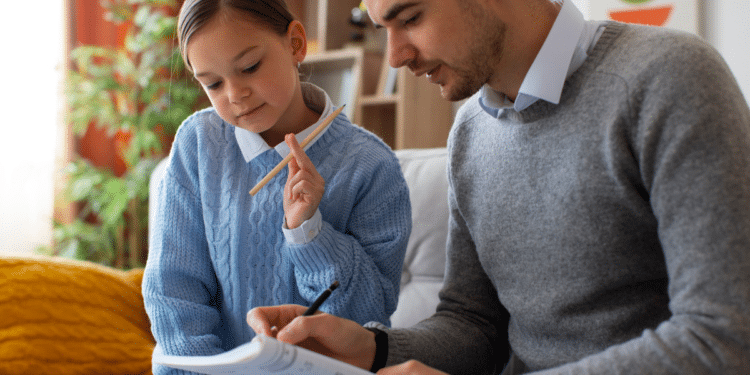 Pai de suéter cinza escreve em caderno enquanto filha de suéter azul segura lápis, sentados no sofá