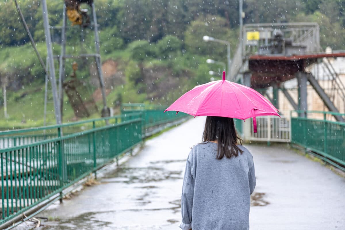 Mulher caminha sobre ponte metálica protegida por um guarda-chuva rosa durante chuva forte, representando os efeitos do El Niño nas regiões afetadas.