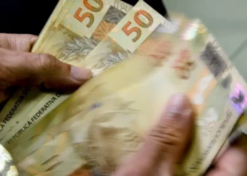 Notas de 50 reais sendo manuseadas, representando os pagamentos do INSS e 13º salário de abril.