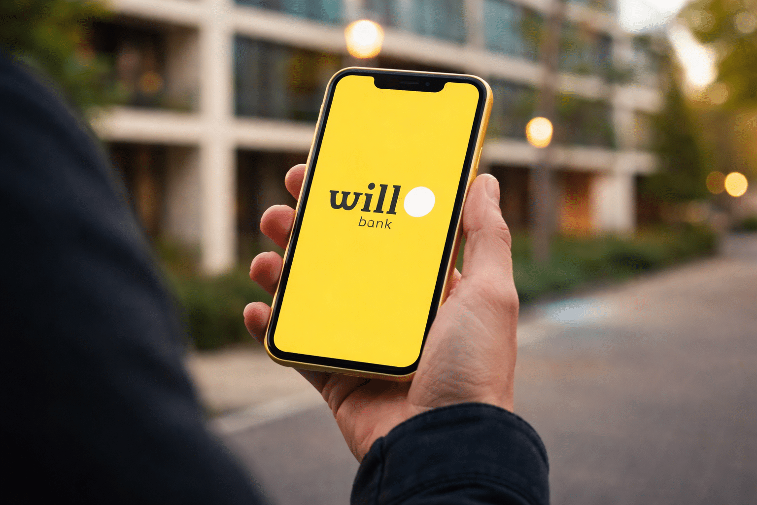 Mão segurando celular com logo do Will Bank na tela amarela, em ambiente urbano externo