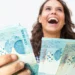 Estudante segurando notas de cem reais representando pagamento do benefício Pé-de-Meia em maio.