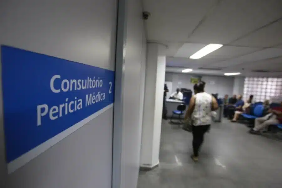 Um consultório de perícia médica do INSS, com o nome 'Consultório Perícia Médica' visível na porta e pessoas aguardando atendimento.