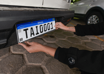 Mãos posicionando placa Mercosul TAI0001 com faixa azul, QR Code e bandeira do Brasil em para-choque traseiro