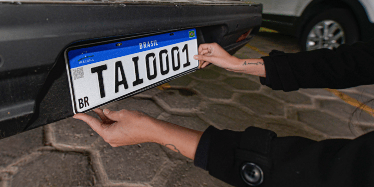 Mãos posicionando placa Mercosul TAI0001 com faixa azul, QR Code e bandeira do Brasil em para-choque traseiro