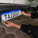 Mãos posicionando placa Mercosul TAI0001 com faixa azul, QR Code e bandeira do Brasil em para-choque traseiro