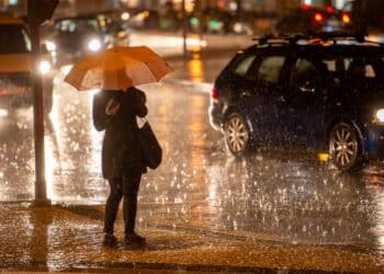 Pessoa com guarda-chuva atravessando rua durante chuva intensa à noite em área urbana.