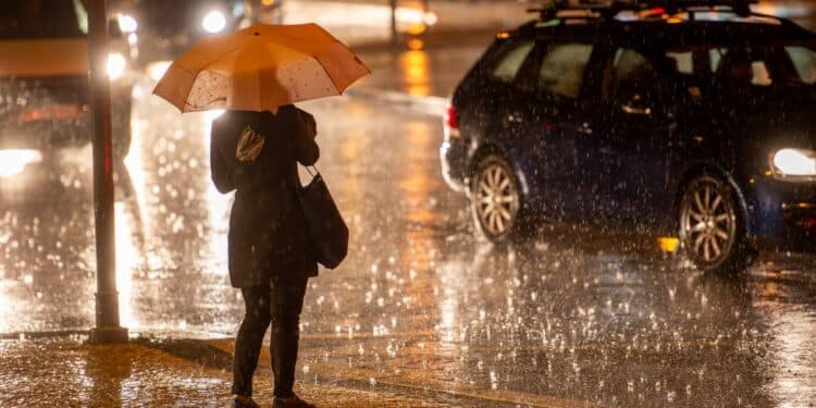 Pessoa com guarda-chuva atravessando rua durante chuva intensa à noite em área urbana.