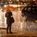 Pessoa com guarda-chuva atravessando rua durante chuva intensa à noite em área urbana.