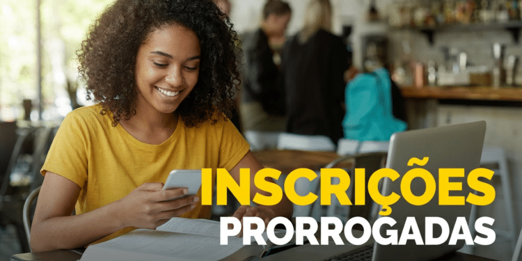 Mulher negra de cabelos cacheados e camiseta amarela segura celular enquanto sorri, com laptop e livro aberto sobre a mesa