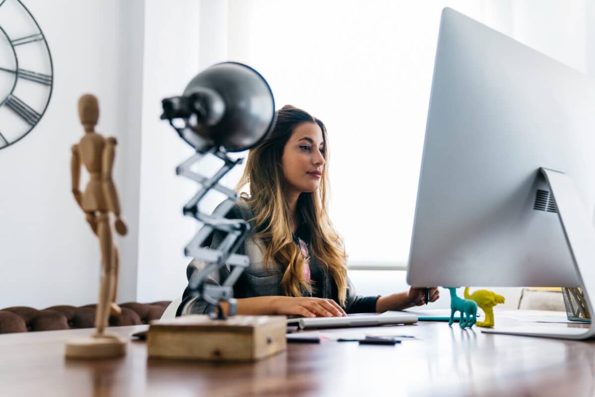 Profissional trabalha concentrada em frente a um computador desktop em ambiente de home office bem iluminado, com luminária e objetos decorativos sobre a mesa.
