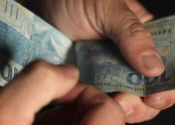 Mãos contando cédulas de R$ 100 do Banco Central do Brasil representando o auxílio mensal do programa Mães de Pernambuco