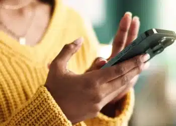 Pessoa digitando em um smartphone, com mãos visíveis, usando um suéter amarelo.