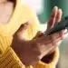 Pessoa digitando em um smartphone, com mãos visíveis, usando um suéter amarelo.