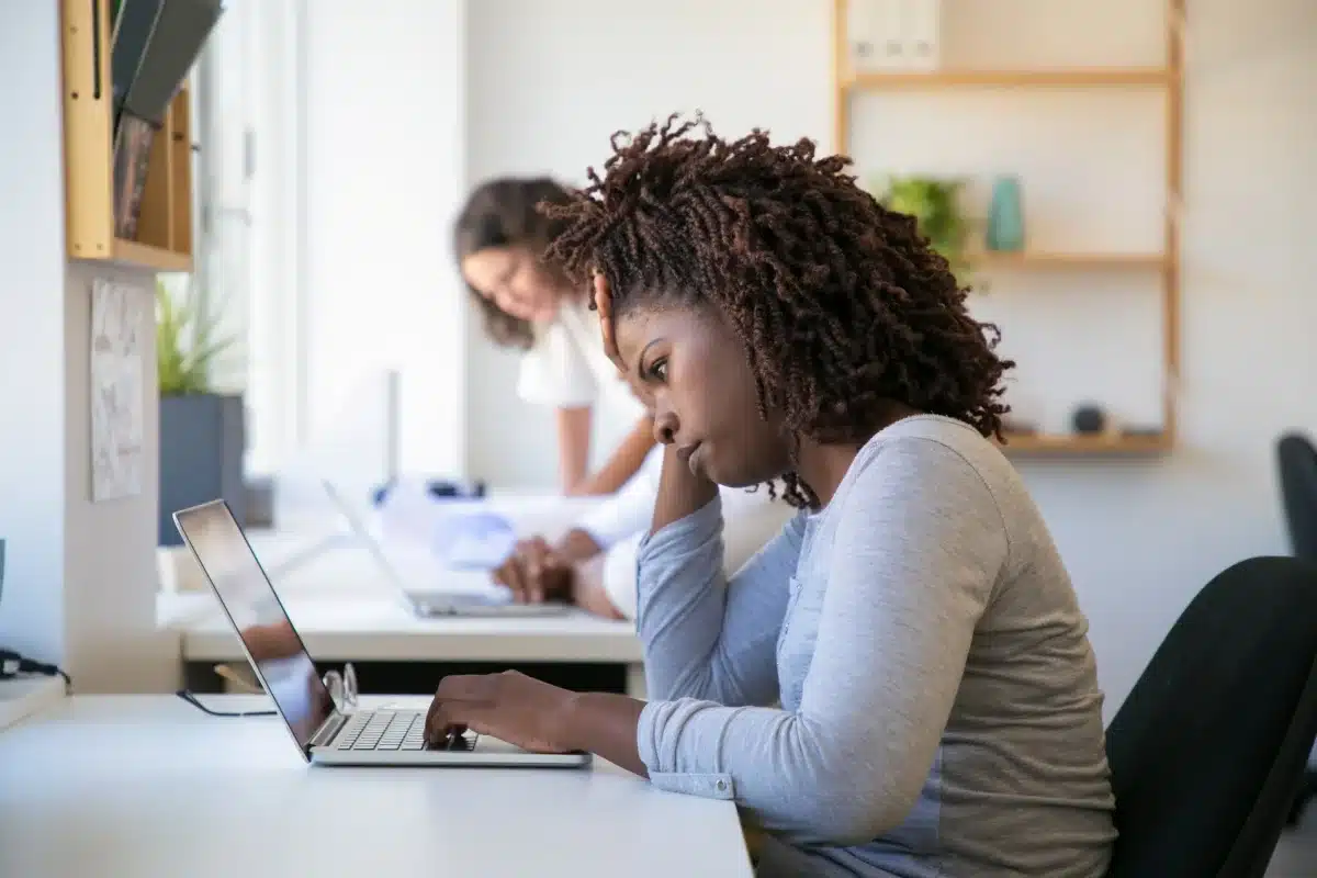 Mulher com cabelo cacheado trabalhando no computador, parecendo cansada, em um ambiente de escritório moderno, representando a importância do fim da escala 6 x 1.