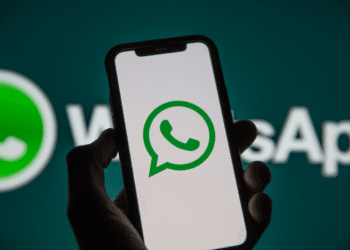 Mão segura smartphone preto com logo do WhatsApp na tela, fundo verde-azulado com marca ampliada do aplicativo