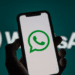 Mão segura smartphone preto com logo do WhatsApp na tela, fundo verde-azulado com marca ampliada do aplicativo