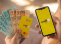 Mãos segurando cartão amarelo Will Bank Mastercard, notas de R$50 e R$100, e celular com app Will Bank aberto
