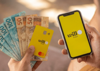 Mãos segurando cartão amarelo Will Bank Mastercard, notas de R$50 e R$100, e celular com app Will Bank aberto